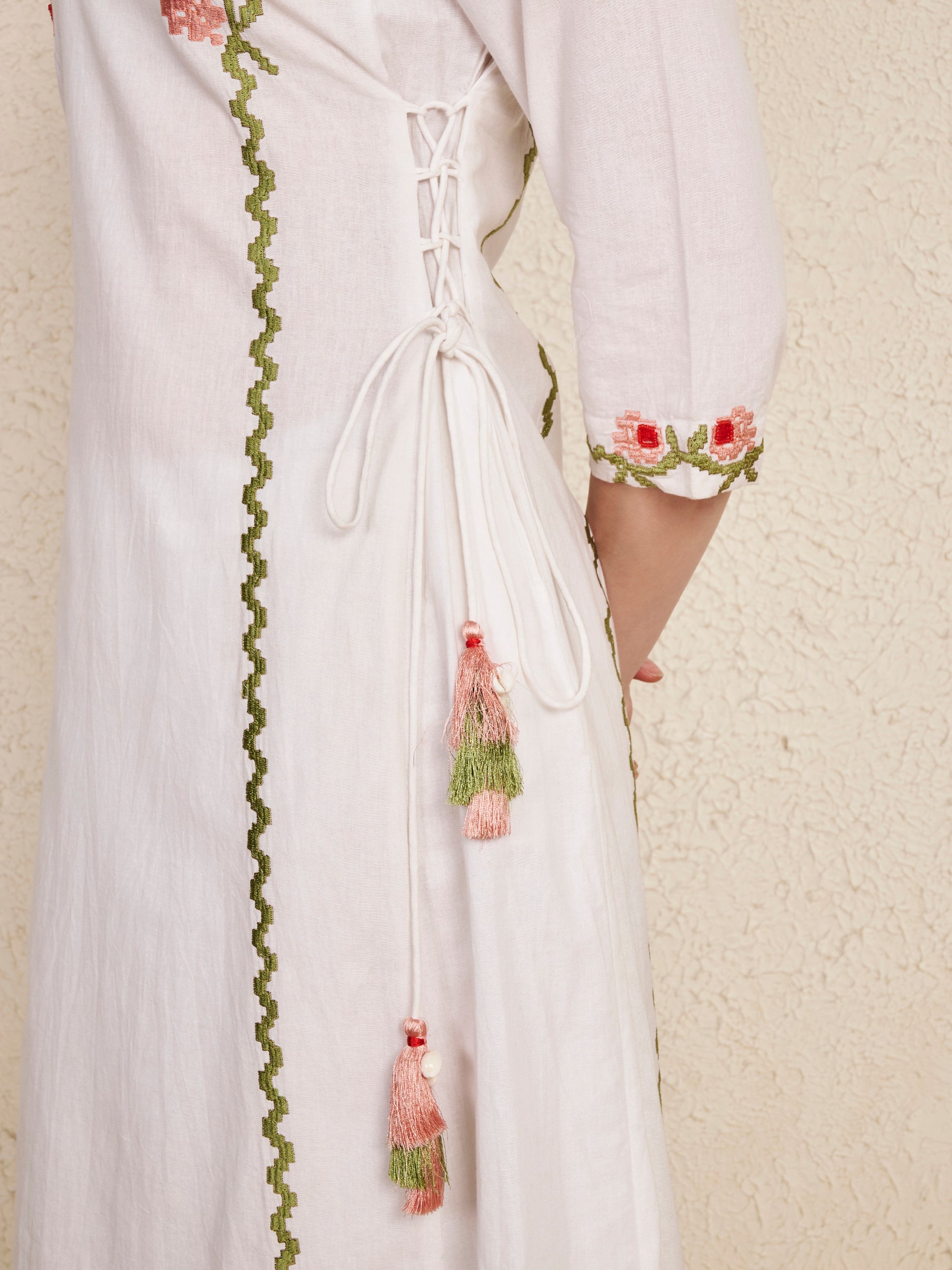 Close-up of a white or off-white embroidered dress or kurta with green zigzag trim, pink floral embroidery, lace-up back detail, and hanging pink and green tassels, shown against a textured beige wall.