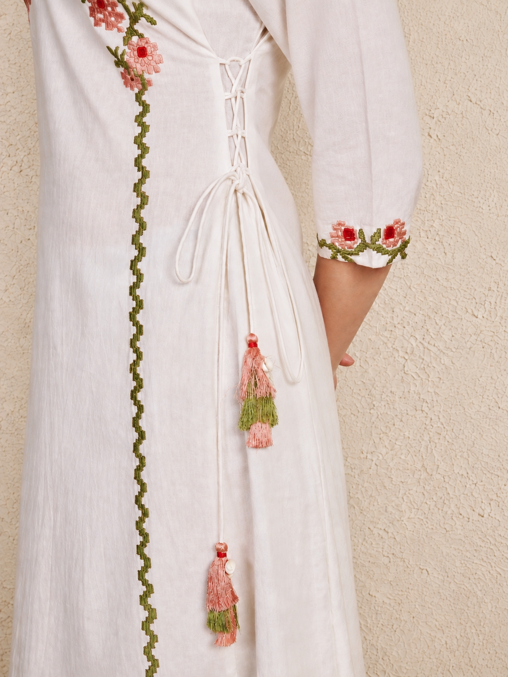 A close-up of a white or off-white embroidered dress with a lace-up back, pink and green floral sleeve embroidery, and dangling pink and green tassels, shown against a textured beige wall.