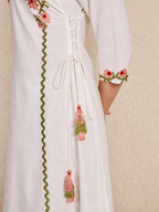 A close-up of a white or off-white embroidered dress with a lace-up back, pink and green floral sleeve embroidery, and dangling pink and green tassels, shown against a textured beige wall.